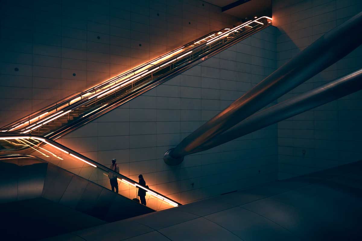 Ambiente futurista contendo dois lances de escadas rolantes com duas pessoas subindo. O corrimão das escadas possui iluminação morna por todo o seu percurso.